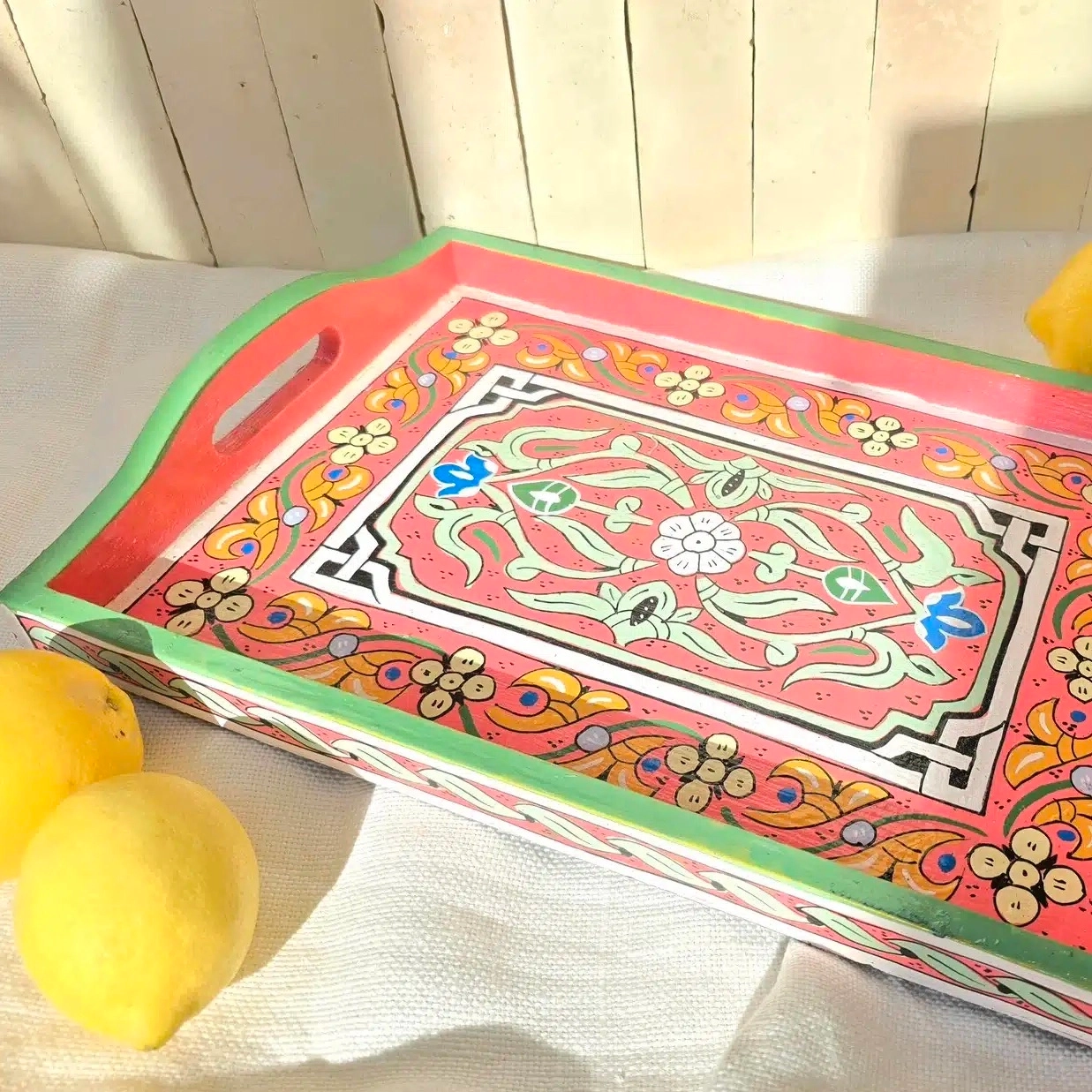 Plateau en bois de cèdre entièrement peint à la main dans un petit atelier à Marrakech.
Chaque pièce demande entre 8 et 9 heures de travail minutieux. Nous avons eu la chance de rencontrer un jeune artisan passionné qui a repris cet atelier. Il a la conviction que l’artisanat doit absolument se transmettre et être valorisé pour perdurer. Il s’engage pour que ces savoir-faire ne soient pas bradés, afin que les artisans puissent en vivre dignement.
Le bois choisi pour la confection de ce plateau est le bois de cèdre, sélectionné pour sa solidité et sa légèreté. L’artisan travaille ensuite au pinceau de calligraphie en poil d’âne, idéal pour un tracé précis et une application fluide de la peinture. Il dessine les motifs sans pochoir ni tracé préalable, avec beaucoup de patience, ajustant les couleurs à l’oeil.
Ce plateau en bois peint est aussi pratique que joli. Il est facile d’utilisation grâce à ses bords relevés et poignées. Idéal pour y servir thé et autres boissons, il peut aussi servir de décoration simplement posé sur une table ou un buffet avec quelques objets du quotidien. Ses couleurs et ses motifs en font en effet un bel élément à laisser visible, qui n’a pas besoin d’être rangé après usage.
Dimensions: Celui-ci mesure 34 x 24 cm