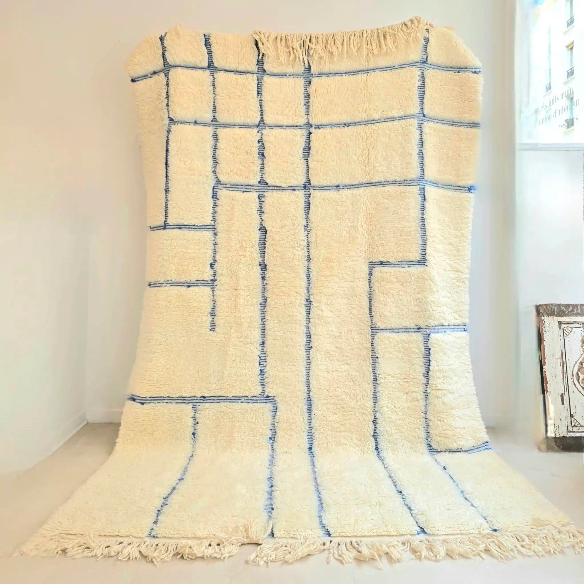 Tapis Berbère Beni Ouarain blanc cassé aux rayures bleues et motifs en relief, réalisé à la main dans les montagne du Haut Atlas Marocain, selon un savoir faire transmis de génération en génération. Il est en pure laine vierge de mouton.
Apportez du caractère et de la douceur à votre intérieur grâce à ce tapis Beni Ouarain fait main. Avec son blanc cassé et ses rayures bleues irrégulières, il s’intègre parfaitement à une décoration contemporaine ou plus classique. Tissé en laine épaisse, il allie authenticité et confort.
Sa texture dense et moelleuse est en effet idéale pour marcher pieds nus ou pour un espace cosy dans votre pièce à vivre.
Son design à la fois moderne et intemporel ainsi que la qualité supérieure de sa laine en font un pièce qui vous suivra de nombreuses années. Les lignes bleues apportent une touche de fraîcheur qui rappelle les maisons de vacances méditerranéennes. Elles sont obtenues grâce à des techniques traditionnelles de teinture artisanale, soulignant l’authenticité du tapis.
Placez-le dans un salon pour structurer l’espace et apporter une ambiance chaleureuse ou dans une chambre pour un effet cocooning. Dans une entrée, il devient un élément décoratif fort qui donne le ton dès l’arrivée.
Dimensions : Il mesure 265 x 160 cm environ