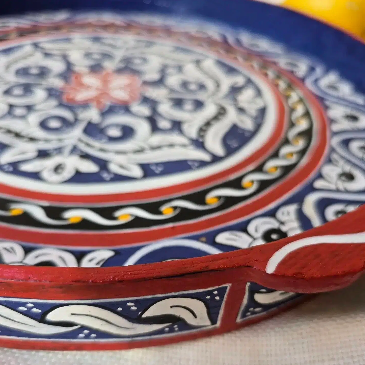Plateau Zouak rond en bois de cèdre entièrement peint à la main dans un petit atelier à Marrakech.
Chaque pièce demande entre 8 et 9 heures de travail minutieux. Nous avons eu la chance de rencontrer un jeune artisan passionné qui a repris cet atelier. Il a la conviction que l’artisanat doit absolument se transmettre et être valorisé pour perdurer. Il s’engage pour que ces savoir-faire ne soient pas bradés, afin que les artisans puissent en vivre dignement.
Le bois choisi pour la confection de ce plateau est le bois de cèdre, sélectionné pour sa solidité et sa légèreté. L’artisan travaille ensuite au pinceau de calligraphie en poil d’âne, idéal pour un tracé précis et une application fluide de la peinture. Il dessine les motifs sans pochoir ni tracé préalable, avec beaucoup de patience, ajustant les couleurs à l’oeil.
Ce plateau en bois peint est aussi pratique que joli. Il est facile d’utilisation grâce à ses bords relevés et poignées. Idéal pour y servir thé et autres boissons, il peut aussi servir de décoration simplement posé sur une table ou un buffet avec quelques objets du quotidien. Ses couleurs et ses motifs en font en effet un bel élément à laisser visible, qui n’a pas besoin d’être rangé après usage.
Dimensions: Celui-ci mesure 35 cm de diamètre