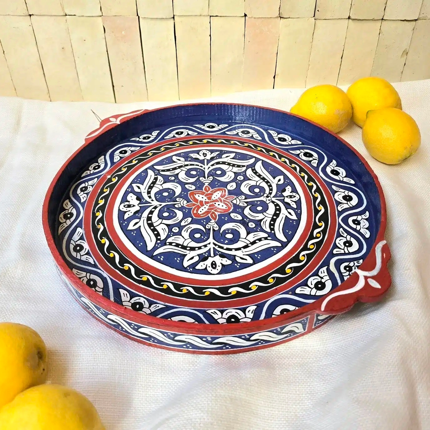 Plateau Zouak rond en bois de cèdre entièrement peint à la main dans un petit atelier à Marrakech.
Chaque pièce demande entre 8 et 9 heures de travail minutieux. Nous avons eu la chance de rencontrer un jeune artisan passionné qui a repris cet atelier. Il a la conviction que l’artisanat doit absolument se transmettre et être valorisé pour perdurer. Il s’engage pour que ces savoir-faire ne soient pas bradés, afin que les artisans puissent en vivre dignement.
Le bois choisi pour la confection de ce plateau est le bois de cèdre, sélectionné pour sa solidité et sa légèreté. L’artisan travaille ensuite au pinceau de calligraphie en poil d’âne, idéal pour un tracé précis et une application fluide de la peinture. Il dessine les motifs sans pochoir ni tracé préalable, avec beaucoup de patience, ajustant les couleurs à l’oeil.
Ce plateau en bois peint est aussi pratique que joli. Il est facile d’utilisation grâce à ses bords relevés et poignées. Idéal pour y servir thé et autres boissons, il peut aussi servir de décoration simplement posé sur une table ou un buffet avec quelques objets du quotidien. Ses couleurs et ses motifs en font en effet un bel élément à laisser visible, qui n’a pas besoin d’être rangé après usage.
Dimensions: Celui-ci mesure 35 cm de diamètre