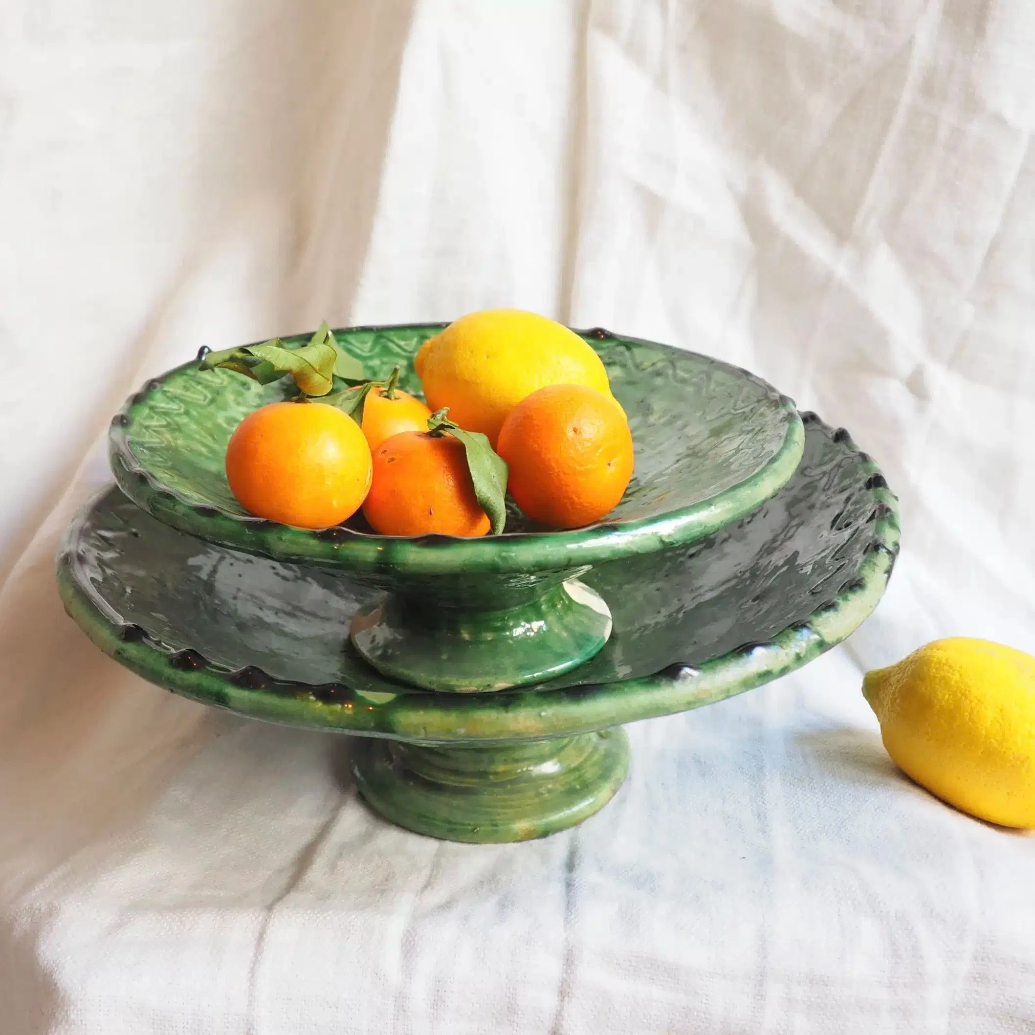 Plat à pied Marocain Tamegroute émaillé vert entièrement réalisé à la main. Ajoutez une touche authentique et bohème chic à votre décoration d’intérieur grâce à cette vaisselle traditionnelle Marocaine unique aux matériaux nobles et naturels
Utilisez en cuisine tous les jours pour vos salades, apéritifs, en coupes à desserts etc… Détournez les en vide-poche, porte-savons…
Ces poteries portent le nom du village où elles sont réalisées: Tamegroute. Seulement quelques familles les fabriquent aujourd’hui, selon un savoir-faire ancestral. Les artisans façonnent chaque pièce à la main et les enduisent ensuite d’un alliage de manganèse et de cuivre. C’est la cuisson à très haute température dans des fours anciens qui apporte leur couleur verte typique, qui varie d’intensité à chaque pièce.
Veuillez noter qu’aucune pièce n’est parfaite: fissures, coulures, bosses, asymétries, etc font partie intégrante des poteries de Tamegroute. La couleur verte diffère à chaque objet, de clair à foncé, et peut parfois ne pas recouvrir la pièce en totalité, laissant ainsi découvrir l’argile.
Ils trouveront leur place dans toutes les pièces de la maison. Disposez les seuls ou en accumulation, en jouant avec leurs différentes tailles, sur une table dans le salon, dans une chambre, sur un buffet, dans une salle de bain…
Ne passent pas au lave-vaisselle
Diamètres : Ils mesurent environ 24 cm et 30 cm de diamètre environ