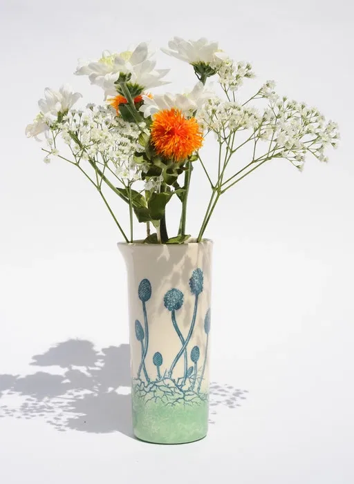Ce vase en céramique évoque un paysage végétal poétique : de délicates tiges surmontées de fleurs graphiques s'élèvent d'une base verte texturée, comme enracinées dans la terre.